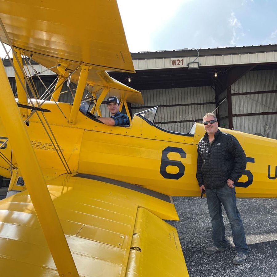 yellow stearman in front of hangar with one man in the cockpit and another on the ground behind the wing