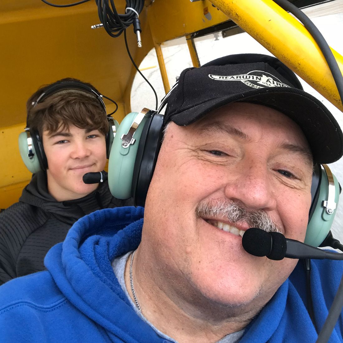 Jim Baker grinning in the cockpit of the Piper Cub. There is a teenage passenger in the back seat.