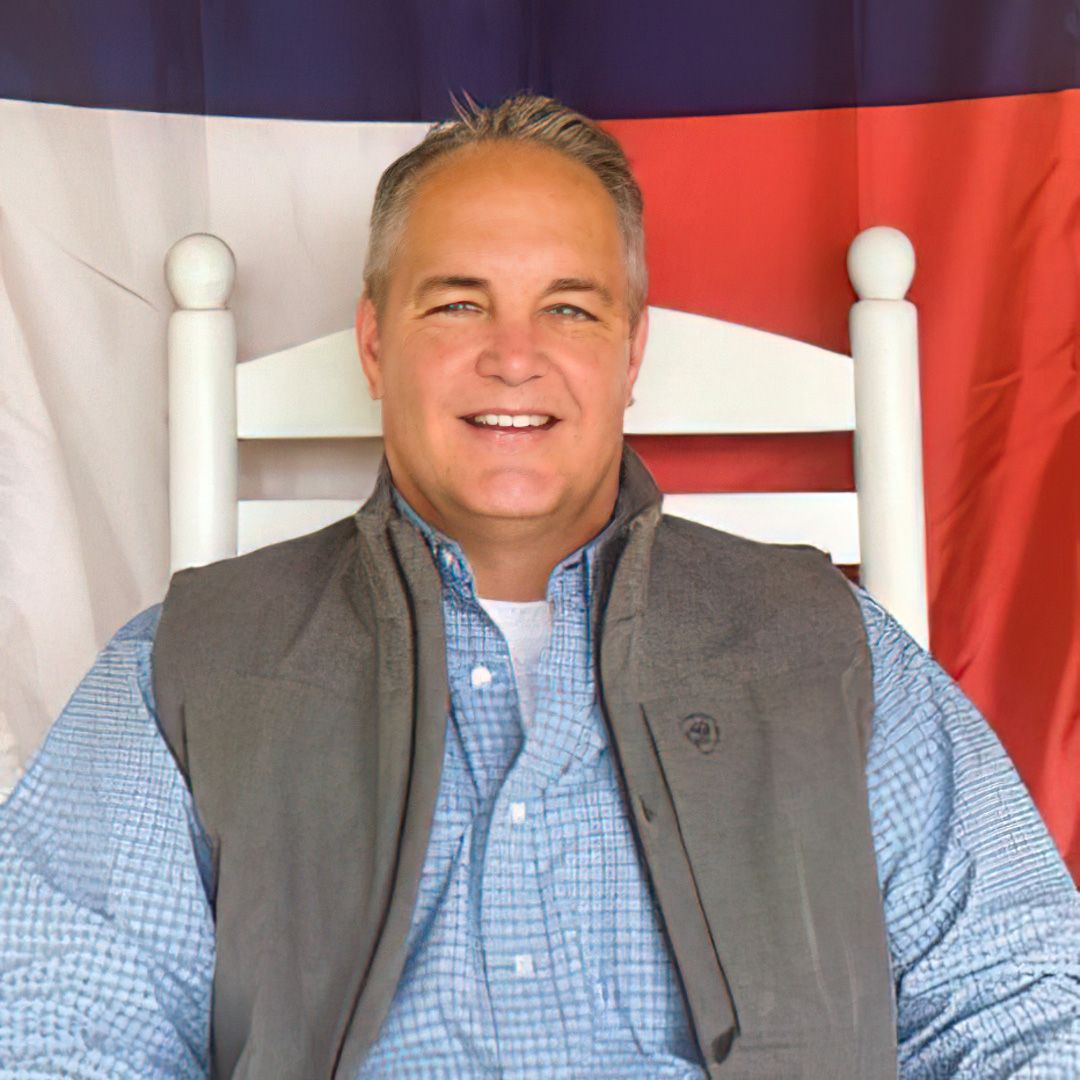 Steven Greenwell in a blue button-down shirt and gray vest sitting in a white chair in front of the Texas flag