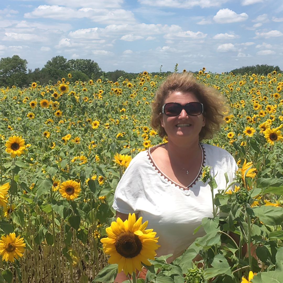 LIsa baker in a short-sleeved white top and big sunglasses standing in a vast field of sunflowers