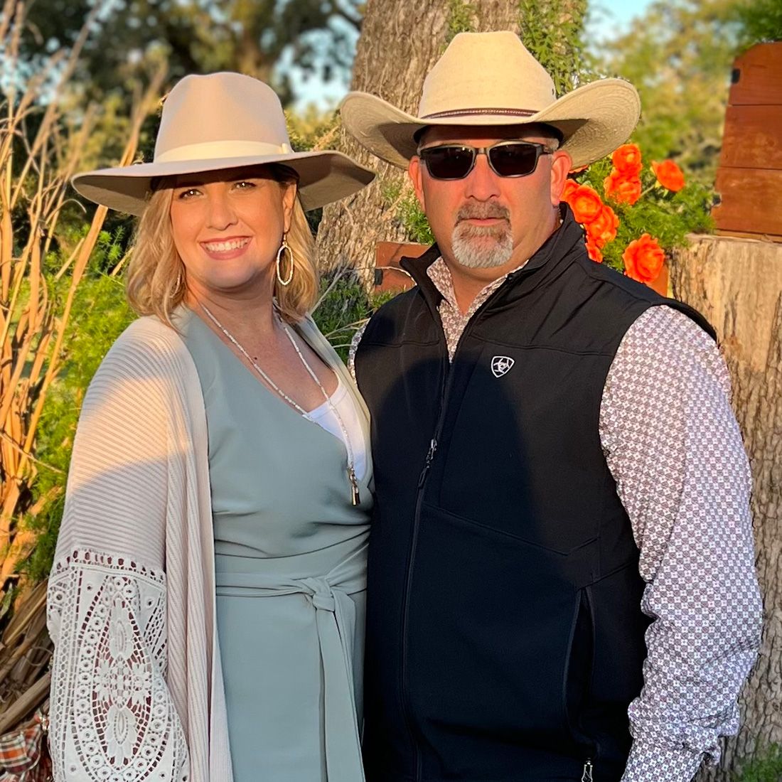 Kelly and Farrah Jernigan standing together in a natural setting. kelly is wearing a black vest and white cowboy hat. farrah is wearing pale green and a beige sweater and light hat.